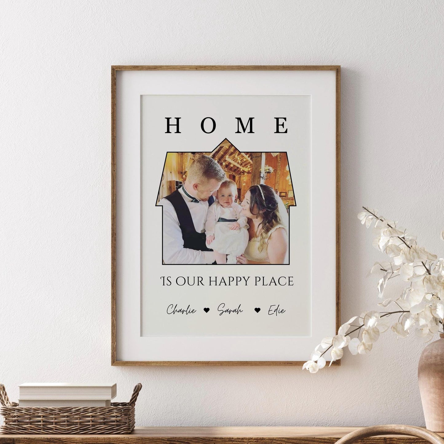 A framed print featuring a a photo set within the outline of a house. Above the image, the word 'HOME' is written in bold, capital letters. Below the house-shaped frame, the phrase 'IS OUR HAPPY PLACE' is printed in a stylish font. The bottom of the print includes the names 'Charlie, Sarah, Effie, Ralphie' separated by small heart symbols. The frame is black, and the print has a clean, minimalist design.
