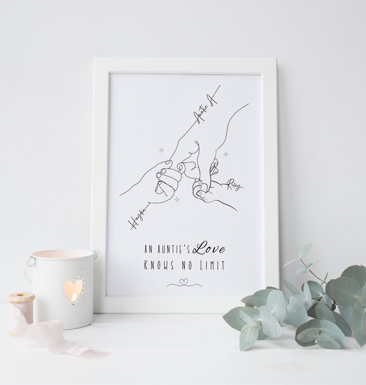 A framed minimalist line art print hanging featuring an illustration of an aunt holding the hands of her nieces or nephews. The simple black line drawing shows delicate hands each labeled with names and The phrase “An Aunty’s Love Knows No Limit”